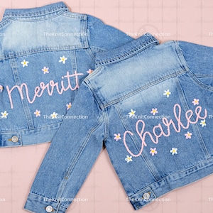 May include: Two denim jackets with embroidered names, "Merritt" and "Charlee", in pink script with white daisy accents.