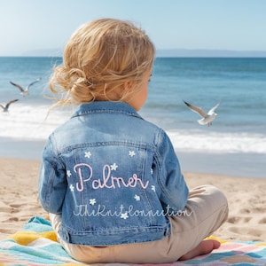 May include: A blue denim jacket with white embroidered flowers and the name "Palmer" embroidered in pink on the back. The jacket is worn by a child sitting on a beach blanket.