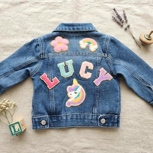 May include: A blue denim jacket featuring the name LUCY in colorful felt letters across the back. The jacket is decorated with embroidered patches of a pink flower, a rainbow, and a unicorn head. It has a collar, long sleeves, and metal buttons.