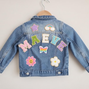 May include: A blue denim jacket featuring the name MAEVE in large, colorful, textured letters across the back. The jacket is decorated with various patches, including a purple star, a rainbow, a butterfly, a pink smiley flower, and a white flower.
