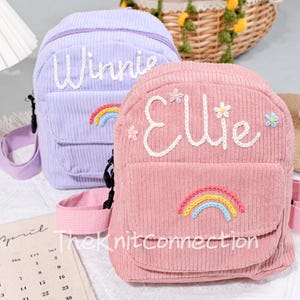 May include: Two small, personalized backpacks with embroidered names, Winnie and Ellie. The backpacks are made of pink and purple corduroy fabric and feature embroidered rainbows and flowers.