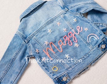 Könnte beinhalten: Eine hellblaue Jeansjacke mit gestickten Blumen und dem Namen "Maggie" in rosa und weißem Garn. Die Jacke hat außerdem einen Regenbogen und eine Sonne.
