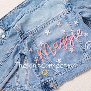 Peut inclure: Une veste en jean bleu clair avec le nom "Maggie" brodé au dos en fil rose et blanc. La broderie comprend également un arc-en-ciel et des fleurs.