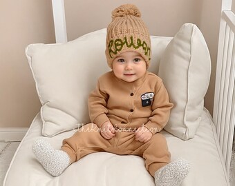 Può includere: Un bambino che indossa un cappello di lana marrone con il nome "Rowan" ricamato in verde. Il bambino indossa una tuta marrone con una piccola tasca sul davanti. Il bambino è seduto su un cuscino bianco.
