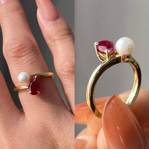 Op de afbeelding: Een gouden ring met een peervormige rode edelsteen en een witte parel. De ring heeft een delicaat ontwerp, met de edelstenen gezet op een dunne band. De ring wordt op een vinger getoond.