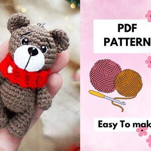 May include: A crochet pattern for a small brown teddy bear wearing a red scarf. The pattern is available as a PDF download and is easy to make. The image also shows two balls of yarn and a crochet hook.