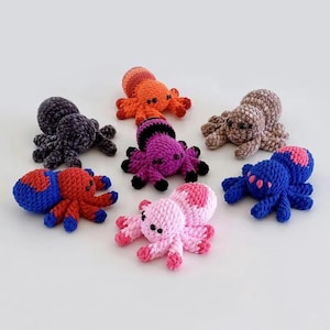May include: Eight crocheted spider toys in various colors, including pink, purple, blue, red, orange, and brown. The spiders have black eyes and are all in a circle on a white background.