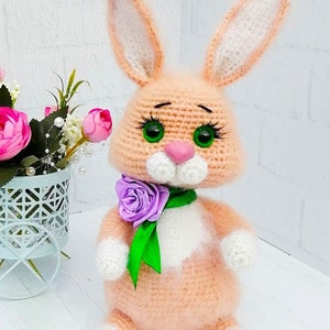 May include: A crocheted plush bunny toy with pink fur, white accents, green eyes, and a purple flower on a green ribbon around its neck.