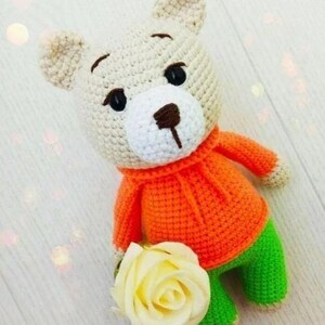May include: A handmade crochet teddy bear with a cream-colored head, white muzzle, and brown nose. The bear wears an orange sweater and green pants, and holds a cream-colored rose. The toy is a charming, handcrafted item.