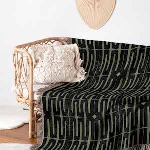 Peut inclure: Une couverture à motifs noirs et vert olive sur un canapé en osier. La couverture présente un motif géométrique avec des franges sur le bord inférieur. Un coussin en macramé et un éventail décoratif sont également visibles.