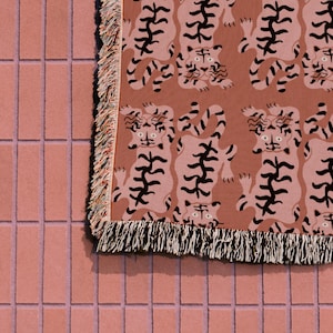 May include: A pink woven throw blanket with a repeating pattern of black tigers. The blanket has a fringe border.