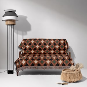 May include: A mid-century modern living room scene featuring a sofa draped with a geometric patterned throw blanket in shades of brown, green, and cream. A black and rattan floor lamp stands to the left, and a woven basket with decorative elements sits on the floor.