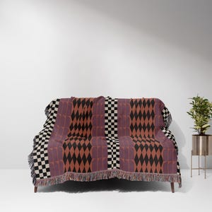 May include: A woven throw blanket with a geometric pattern in shades of red, black, and white. The blanket is draped over a couch with a fringe border.