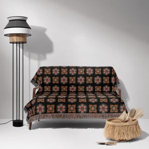 May include: A black sofa with a floral patterned throw blanket. The throw blanket has a fringed border and features a repeating pattern of pink, orange, and green flowers on a black background. The sofa is in a room with a white wall and a black floor lamp.