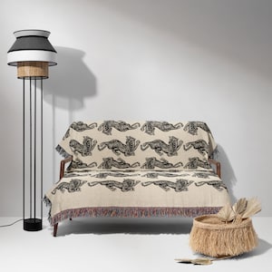 May include: A cream-colored throw blanket with a black tiger print design. The blanket is draped over a small couch with black legs. A black floor lamp with a white and black lampshade is in the background.