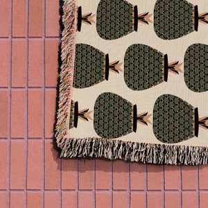 Può includere: Una coperta in tessuto intrecciato con bordo a frangia presenta un motivo ripetuto di ananas verdi su uno sfondo beige.