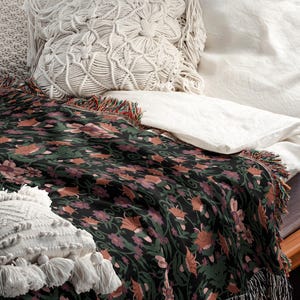 Puede incluir: Una manta de tiro negra y verde con un patrón floral y flecos. La manta está drapeada sobre una cama blanca con almohadas blancas.