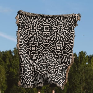 May include: A black and white woven throw blanket with a wavy, abstract pattern. The blanket is being held up in front of a person standing in a grassy field.