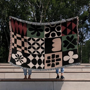 May include: Two people are holding up a woven throw blanket with a geometric pattern in black, white, green, pink, and red. The blanket has a fringe border.