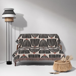 Puede incluir: Una manta de tiro con estampado en blanco y negro con un borde de flecos drapeada sobre un sofá marrón. La manta presenta un diseño geométrico de flores estilizadas. Una lámpara de pie negra con una pantalla blanca está en el fondo.
