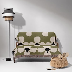 May include: A modern living room scene featuring a green and cream floral patterned throw blanket draped over a wooden sofa. A black and white floor lamp stands to the left, and a woven basket with decorative items sits on the floor.