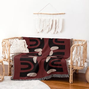May include: A burgundy throw blanket with a black and beige abstract design is draped over a wicker sofa. The blanket has a fringed edge. A decorative wall hanging and a woven pillow are also visible.