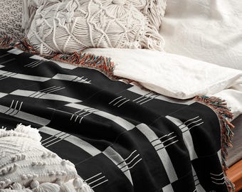 May include: A black and white geometric patterned throw blanket with a fringed edge. The blanket is draped over a bed with white linens and decorative pillows. The pillows are white and have textured designs.