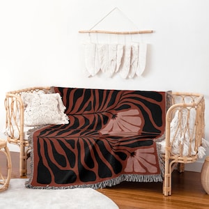 May include: A throw blanket with a floral pattern in black and rust colours. The blanket is draped over a wicker sofa, with a decorative pillow and a macrame wall hanging in the background. The blanket has a fringed edge. The sofa is made of wicker.