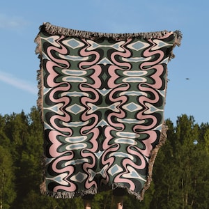 May include: A patterned blanket with a repeating design of pink, black, and green swirls, and light blue diamond shapes. The blanket has a fringed edge and is held up against a blue sky. The blanket is made of a soft material.