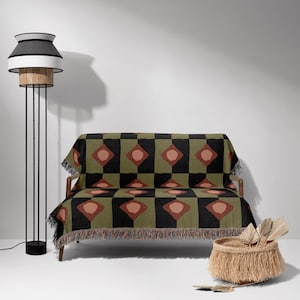 May include: A green, black and brown woven throw blanket with a geometric pattern. The blanket is draped over a small sofa with wooden legs. A wicker basket with a fringe is in the foreground.
