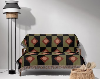 May include: A green, black, and brown woven throw blanket with a geometric pattern. The blanket is draped over a small sofa with wooden legs. A wicker basket with a fringe is in the foreground.