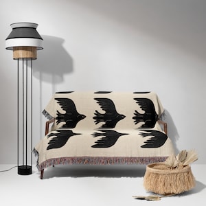 May include: A black and white woven throw blanket with a pattern of flying birds. The blanket is draped over a small couch with wooden legs. A wicker basket with a fringe sits on the floor next to the couch.