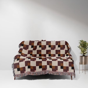 May include: A woven throw blanket with a geometric pattern in shades of brown, black and beige. The blanket is draped over a couch with wooden legs.