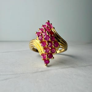 May include: A gold ring featuring a cluster of round, vibrant pinkish-red gemstones arranged in a marquise-shaped setting. The band is crafted from polished yellow gold and displays a smooth, curved design that tapers toward the back of the ring.
