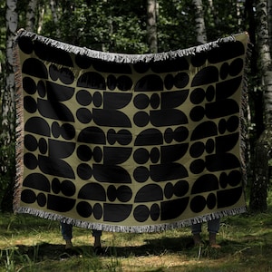 May include: A woven throw blanket with a geometric pattern of black shapes on an olive green background. The blanket has a fringed edge and is held up in front of a forest backdrop.