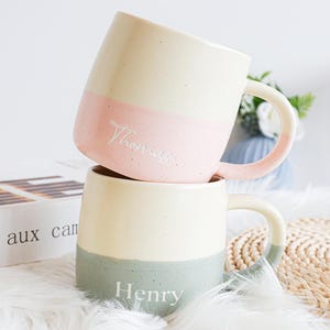 May include: Two ceramic mugs, one stacked on top of the other. The top mug has a pink band with the name "Thomas", the bottom mug has a green band with the name "Henry". Both mugs have a cream upper half and matching handles.