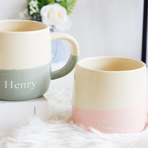 May include: Two ceramic mugs with a two-tone design. One mug is cream and green with the name "Henry" printed on it. The other mug is cream and pink with the name "Thomas" printed on it. Both mugs have a handle.
