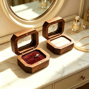 May include: Two octagonal wooden ring boxes with hinged lids and glass inserts. One box features a deep red velvet interior holding a silver ring, while the other has a cream-colored velvet insert. Both lids display etched floral designs.