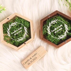 Peut inclure: Deux boîtes à alliances hexagonales en bois avec une base de mousse verte et une inscription blanche. L'inscription indique "Camille & André 08.29.2024". Une clé USB en bois avec la même inscription est également visible.