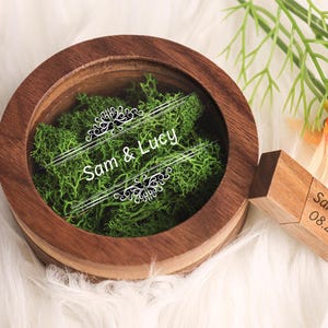May include: A wooden ring box with a glass lid, filled with green moss. The lid is engraved with the names "Sam & Lucy" and a decorative design. A wooden USB drive with the names "Sam & Lucy" and the date "08.29.2024" is attached to the box.