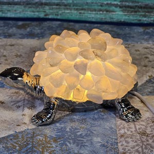 May include: A decorative turtle-shaped lamp with a silver-colored body and legs. The turtle's shell is made of light-colored, irregularly shaped stones that illuminate when the lamp is turned on. The lamp has a power cord.