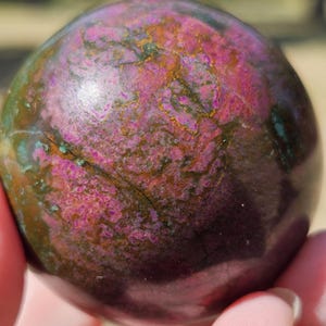 May include: A polished spherical stone featuring a vibrant mix of deep magenta, dark forest green, and earthy brown tones. The surface displays intricate, natural veining and mottled patterns with a smooth, reflective finish across the entire sphere.