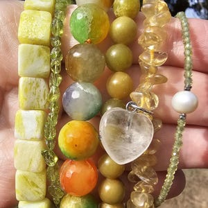 May include: A set of five beaded bracelets featuring yellow square beads, green faceted beads, multicolored round stone beads, yellow chip beads with a clear heart-shaped charm, and a thin green beaded strand with a single white pearl.