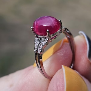 May include: A silver-toned ring featuring a large, round, cabochon-cut deep pink star ruby gemstone set in a four-prong mount. The band is accented with small, clear, sparkling stones arranged in a butterfly-shaped design on each side.