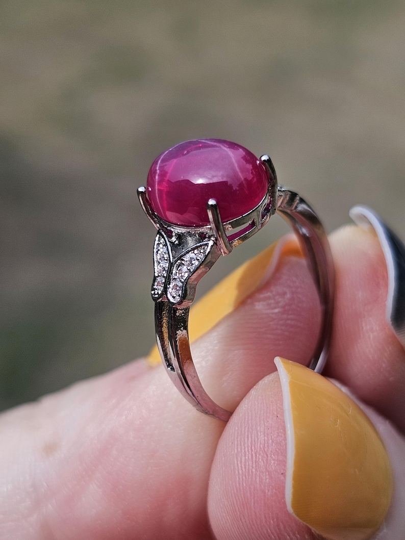 May include: A silver-toned ring featuring a large, round, cabochon-cut deep pink star ruby gemstone set in a four-prong mount. The band is accented with small, clear, sparkling stones arranged in a butterfly-shaped design on each side.