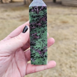 May include: A tall, rectangular zoisite crystal obelisk. The polished stone features a green base with black and magenta inclusions, topped with a gray, pyramidal point. The obelisk is held in a hand against a blurred outdoor background.