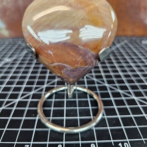 May include: A polished, heart-shaped stone in shades of tan, brown, and burgundy, displayed on a silver metal stand. The stone has natural veining and a glossy finish. The stand features a circular base and two prongs to hold the stone.