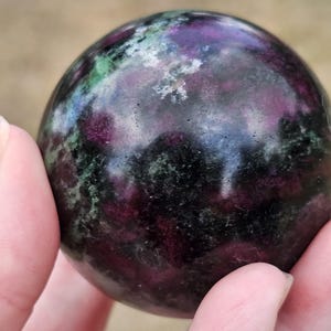 May include: A polished sphere of ruby zoisite, showcasing a blend of deep purple, green, and black hues. The gemstone's surface reflects light, highlighting its unique patterns and textures. This decorative sphere is a natural stone.