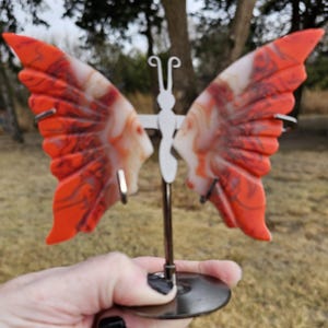 May include: A decorative butterfly sculpture with vibrant orange and white wings, supported by a silver metal stand. The wings have a marbled effect, and the sculpture is displayed outdoors.