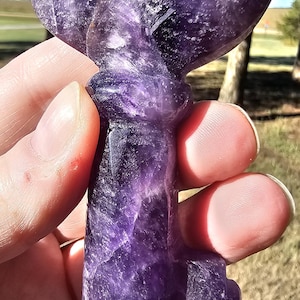 May include: A carved purple amethyst crystal shaped like a skeleton key. The stone features deep violet hues with natural white inclusions and a polished, smooth surface. The head of the key is rounded, and the shaft leads to a notched bit.